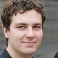 A person with short brown hair, a portion of the ear visible, standing against a neutral background.