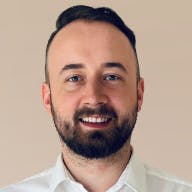 A person wearing a white collared shirt is against a plain beige background. The image focuses on the upper part of the person's body, specifically the neck and shoulders. The person has short, dark hair and a neatly trimmed beard.