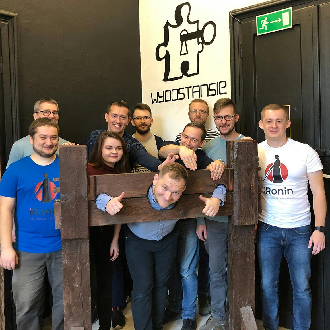 A group of people posing together in an escape room setting, with one person playfully locked in wooden stocks.