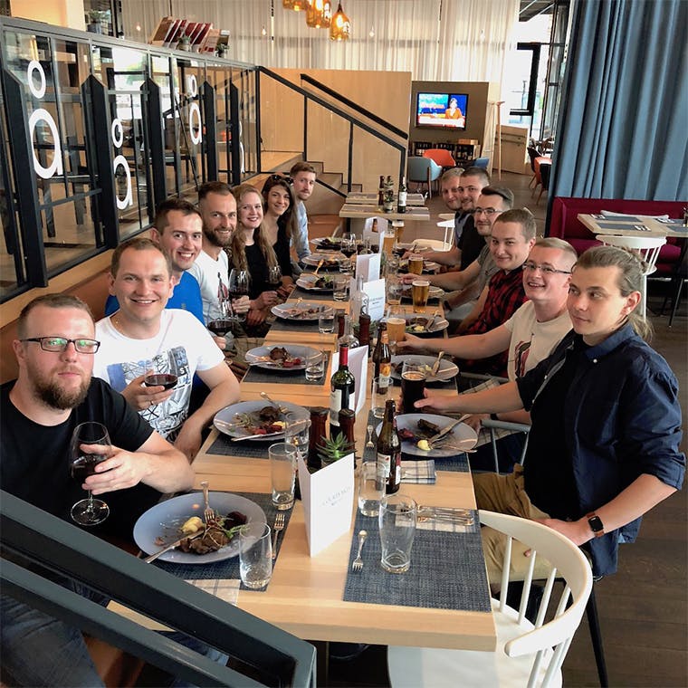 A group of people seated around a long, elegantly arranged dining table in a restaurant. The table is adorned with plates of food, glasses of wine, beer bottles, and other beverages, creating a lively and inviting atmosphere.