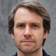 A person with light brown hair is standing against a gray background. The hair is styled with a side part. The person is wearing a dark-colored shirt or jacket.