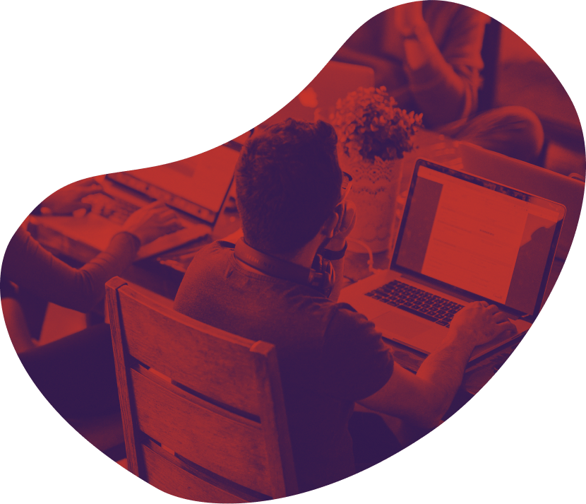 A person sits at a desk using a laptop. Other laptops are partially visible on the desk. The image has a red and purple overlay.