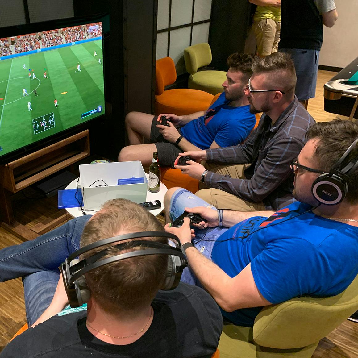 A group of people sitting in a cozy room, engaged in playing a soccer video game displayed on a large television screen. They are holding game controllers and wearing headphones, fully immersed in the gameplay.