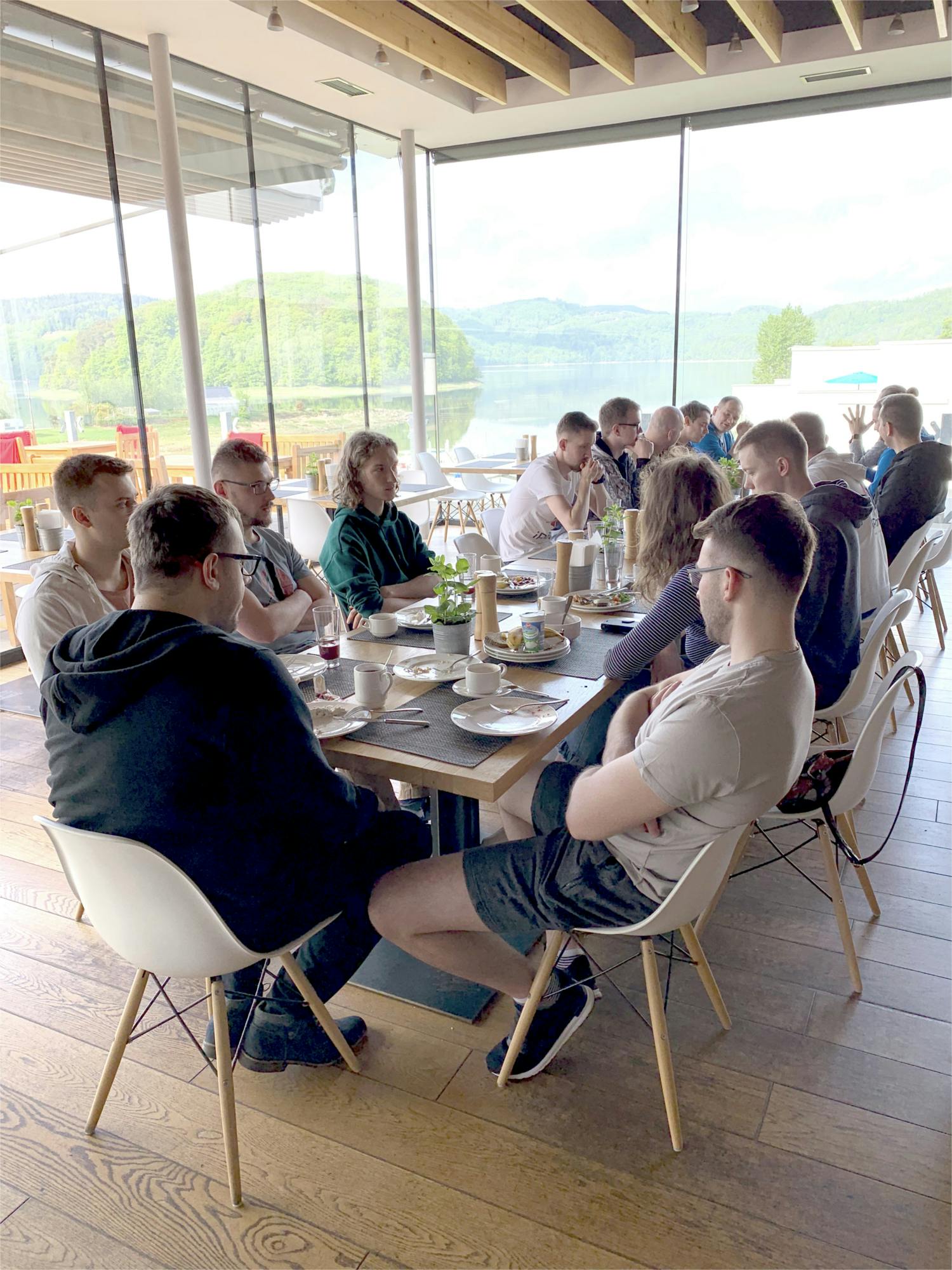 A group of people is sitting around a long wooden table in a modern restaurant with large glass windows overlooking a scenic lake and green hills. The table is set with plates, cups, and condiments, indicating a meal or meeting. The attendees, mostly young adults, are engaged in conversation, some leaning forward while others listen. The atmosphere appears casual and relaxed. Natural light brightens the room, and the wooden floor and ceiling beams add warmth to the setting.